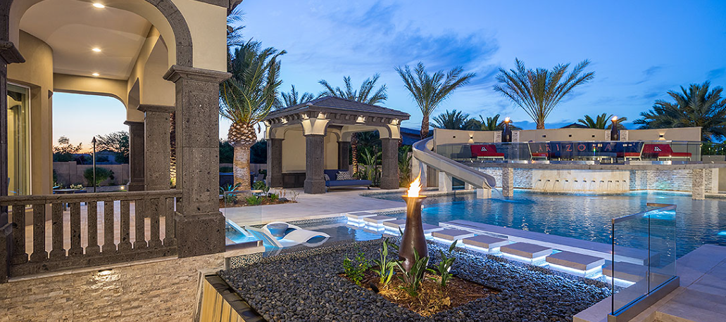Luxury resort-style backyard patio with a pool, waterslide, fire features, and a cabana at dusk.